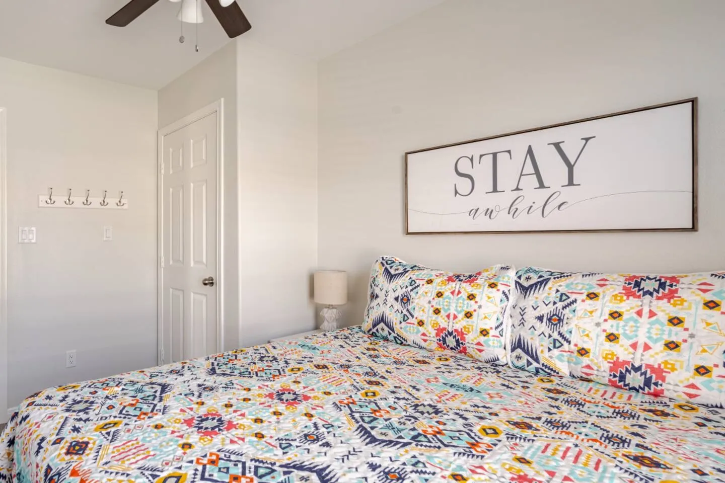 Bedroom corner with a colorful patterned quilt, matching pillows, wall art reading STAY awhile, and an anchor coat rack.