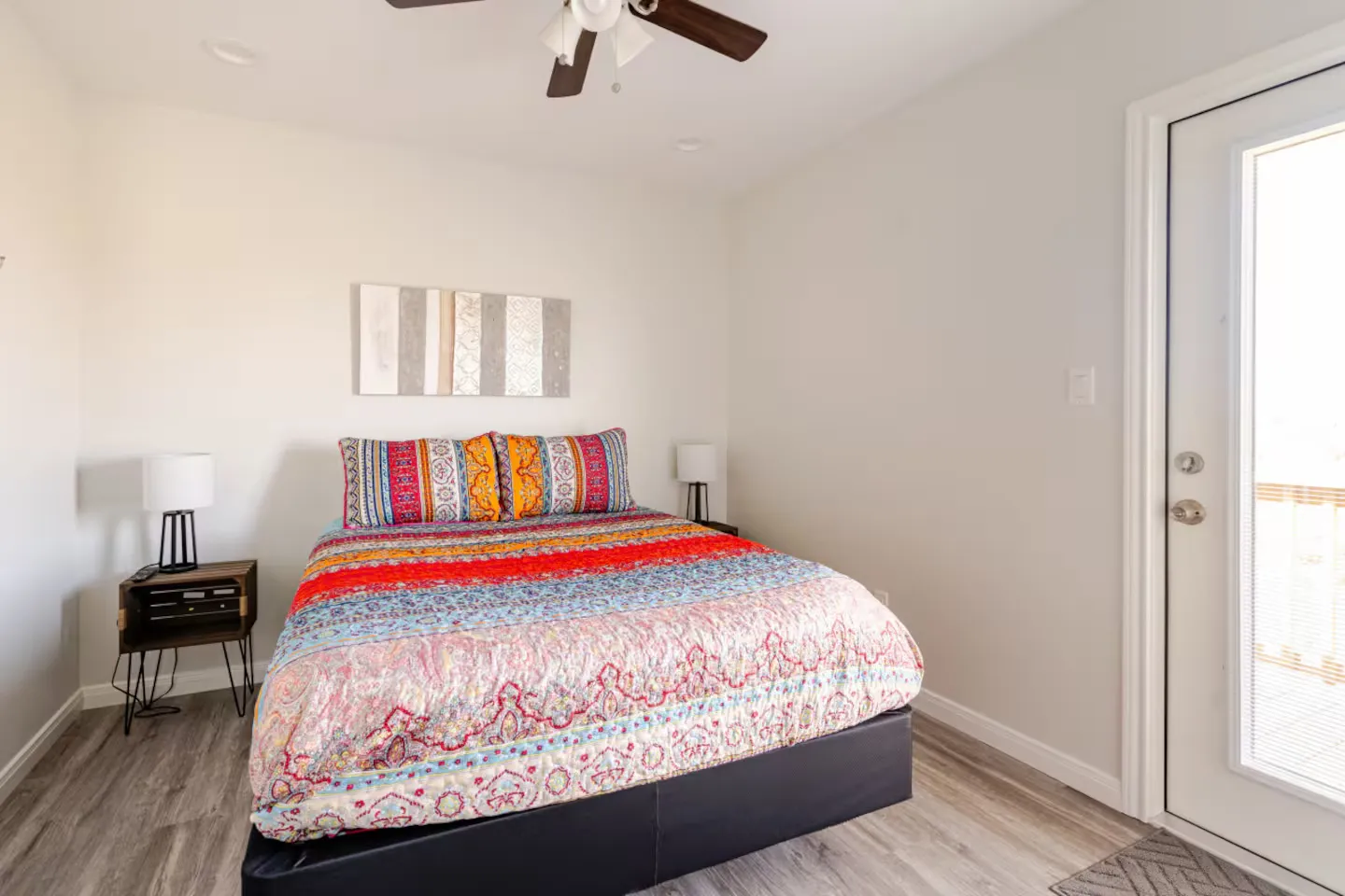 Bedroom with a colorful patterned bedspread, nightstands with lamps, ceiling fan, and an exterior door with glass.