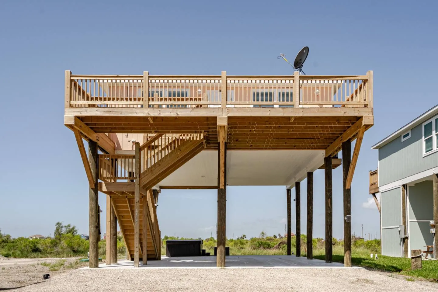 Exterior of an elevated stilt beach home featuring a large wooden deck, wooden staircase, and open covered area beneath the structure.
