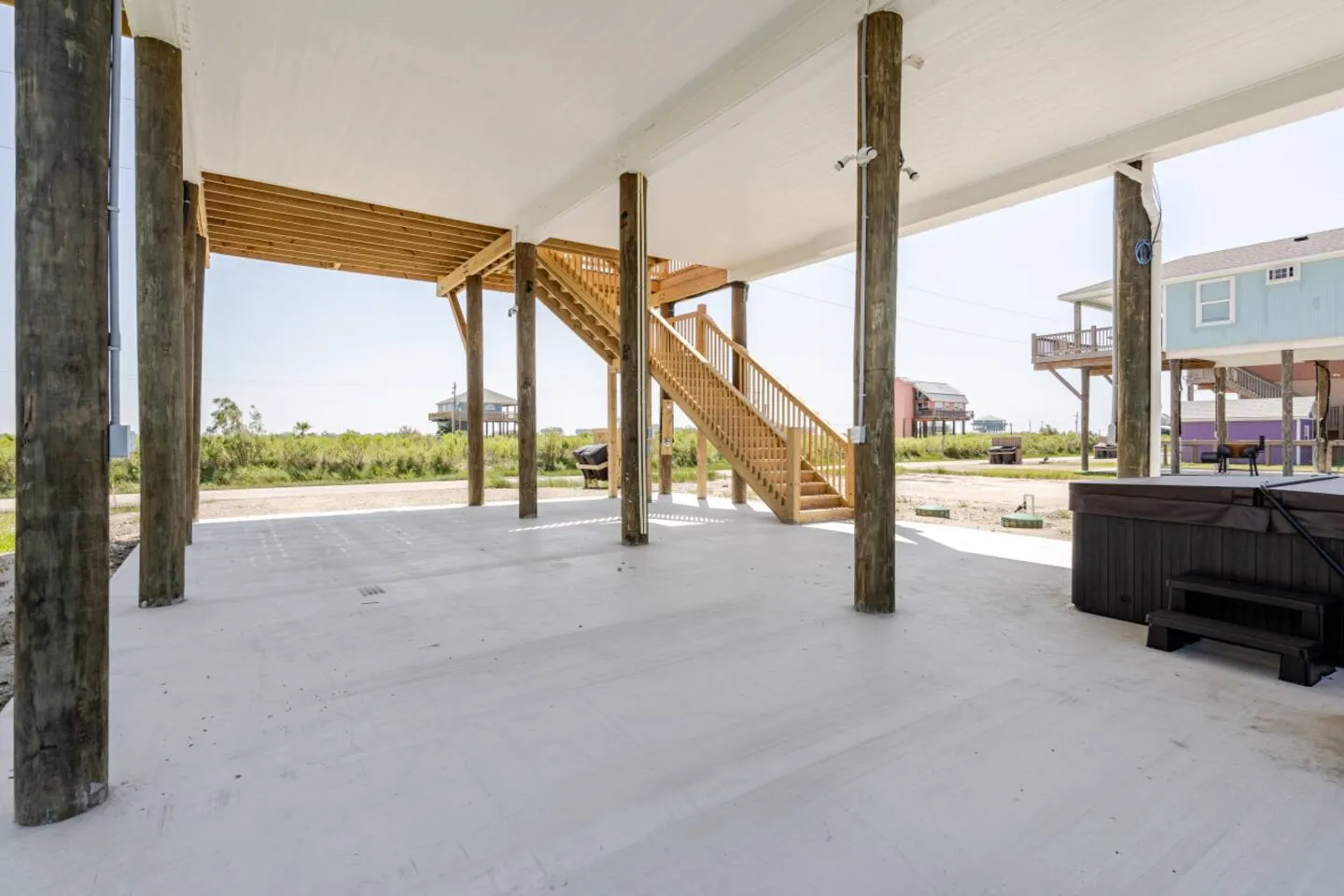 Open ground-level space beneath a stilt home, featuring concrete ground, wooden pilings, exterior stairs, and a covered hot tub.