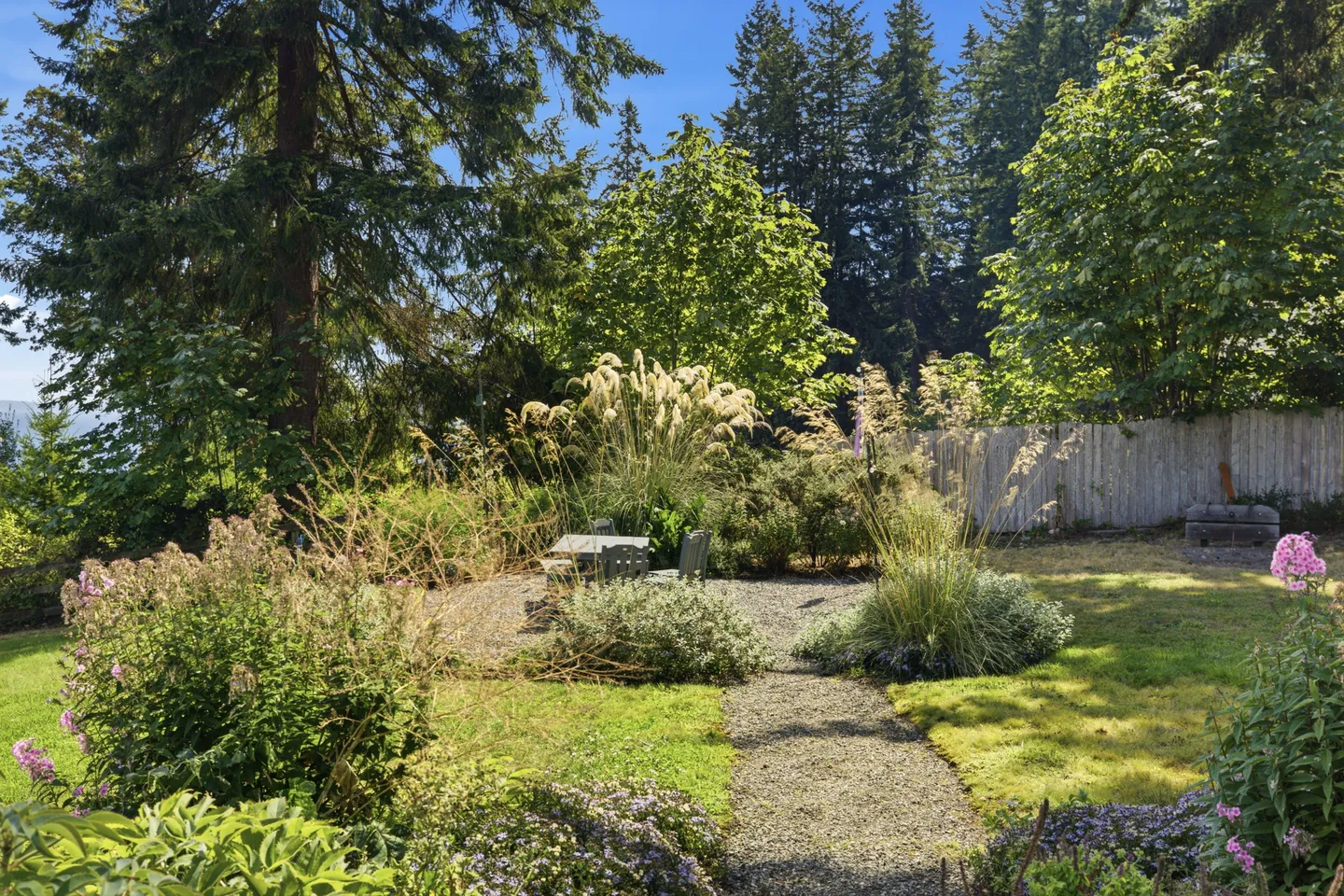 A gravel pathway through a lush grassy backyard leading to an outdoor seating area surrounded by tall grasses, garden plants, and dense evergreen trees.