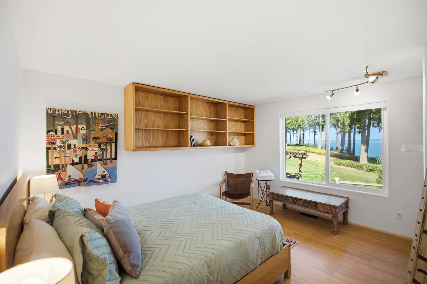 Bedroom with a bed, light wood built-in shelving, wall tapestry, and a large window overlooking trees and water.