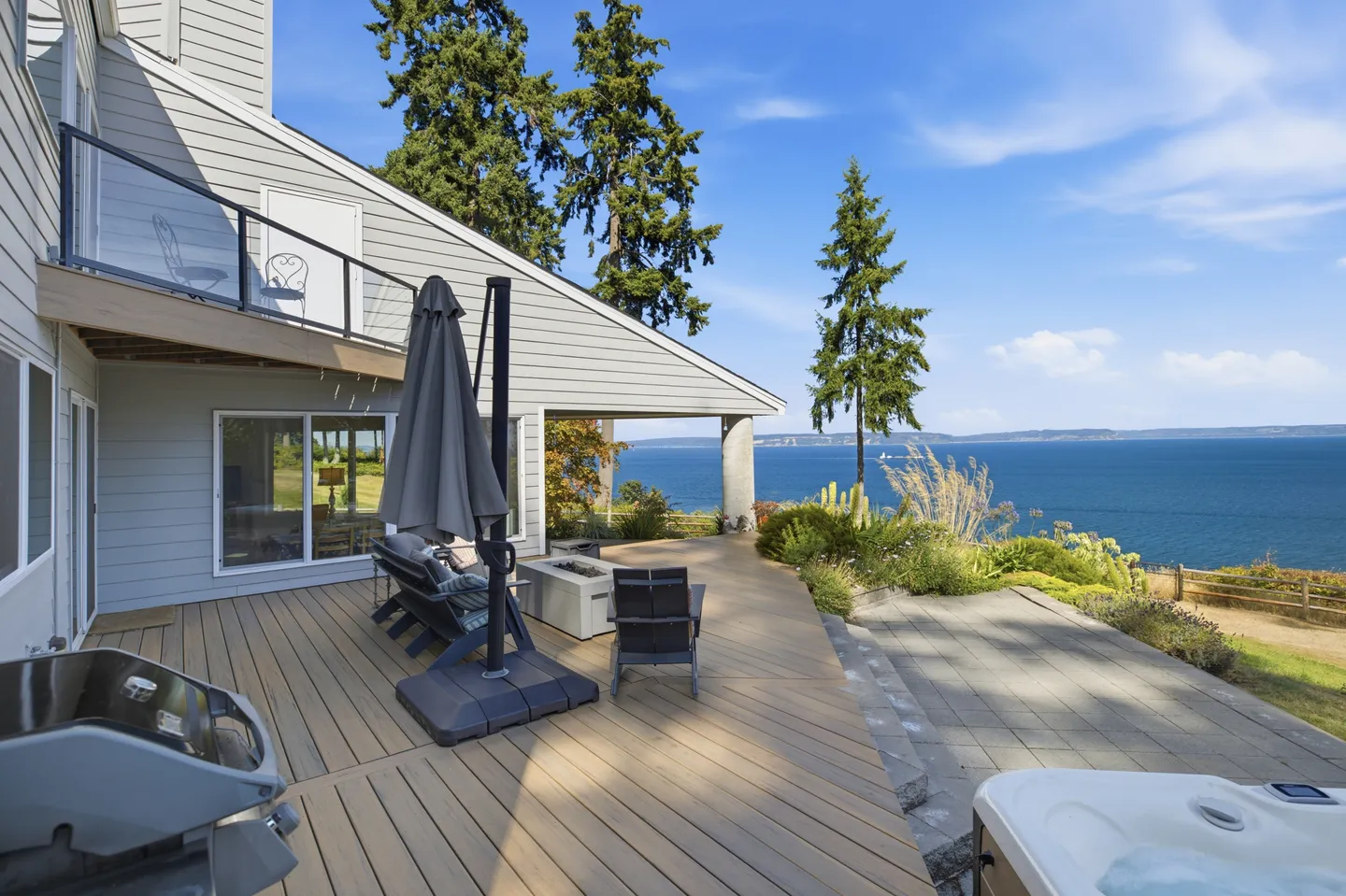 Outdoor deck and patio with seating, a fire table, a grill, a hot tub, and sweeping ocean views under a sunny blue sky.