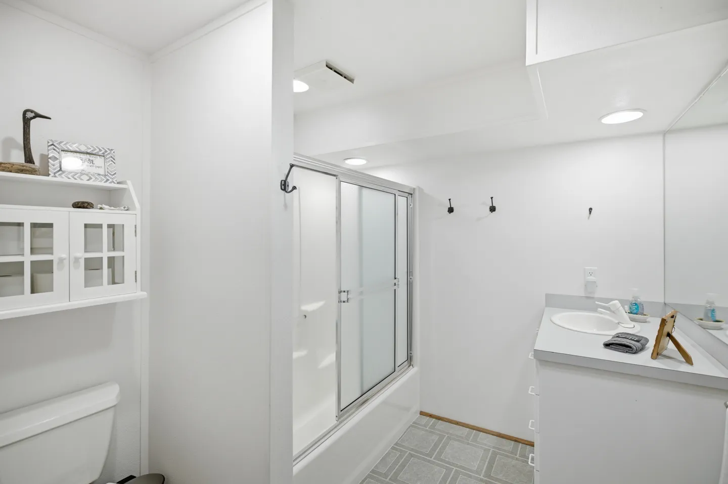 Bathroom with white walls, a vanity with a sink, a toilet with shelving above, and a shower-tub combo with sliding frosted glass doors.
