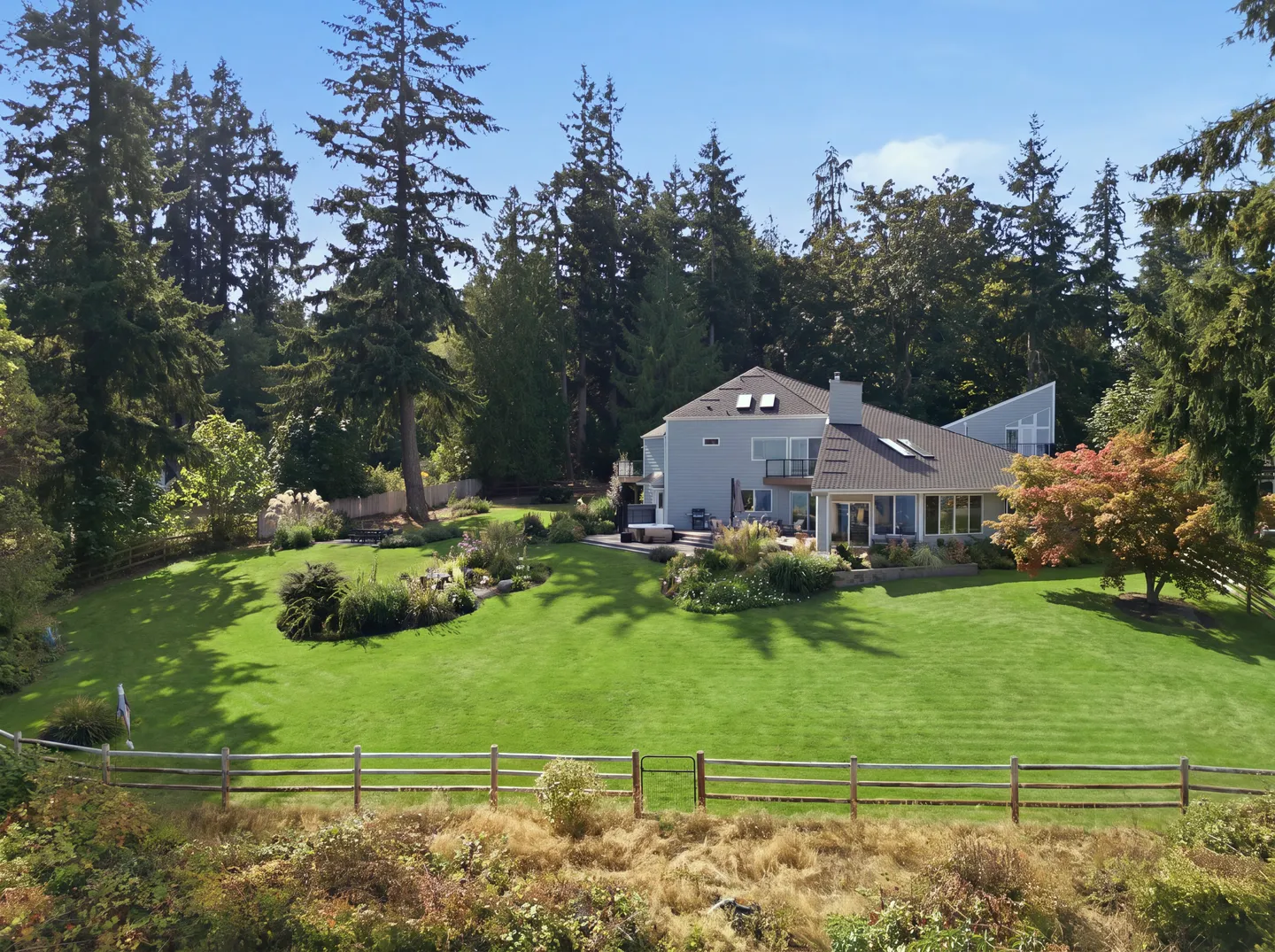 Elevated view of a modern two-story home with a spacious green lawn, landscaped garden beds, and surrounded by tall evergreen trees under a blue sky.