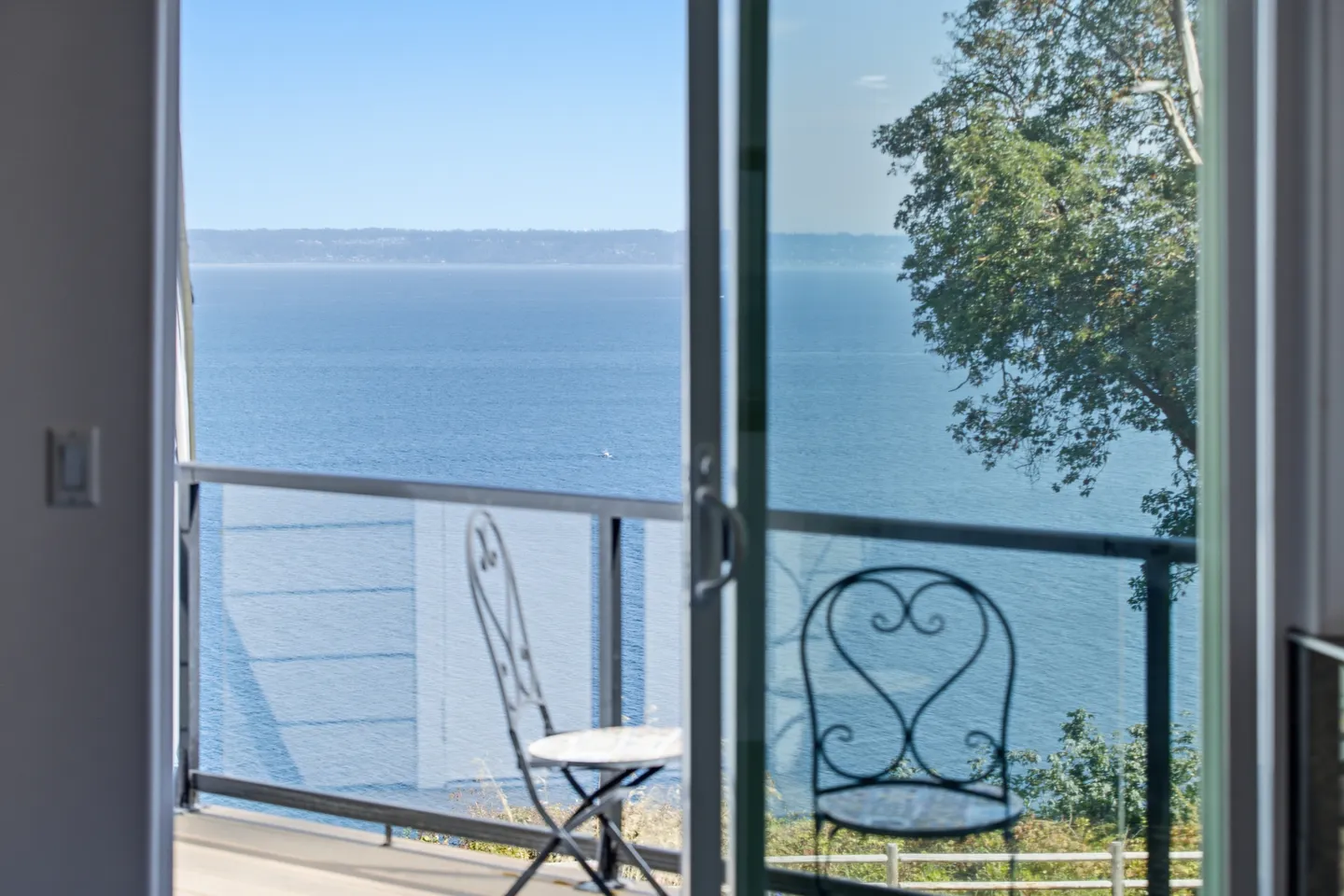 A view of the ocean through a sliding glass door leading to a balcony with glass railings and metal chairs.