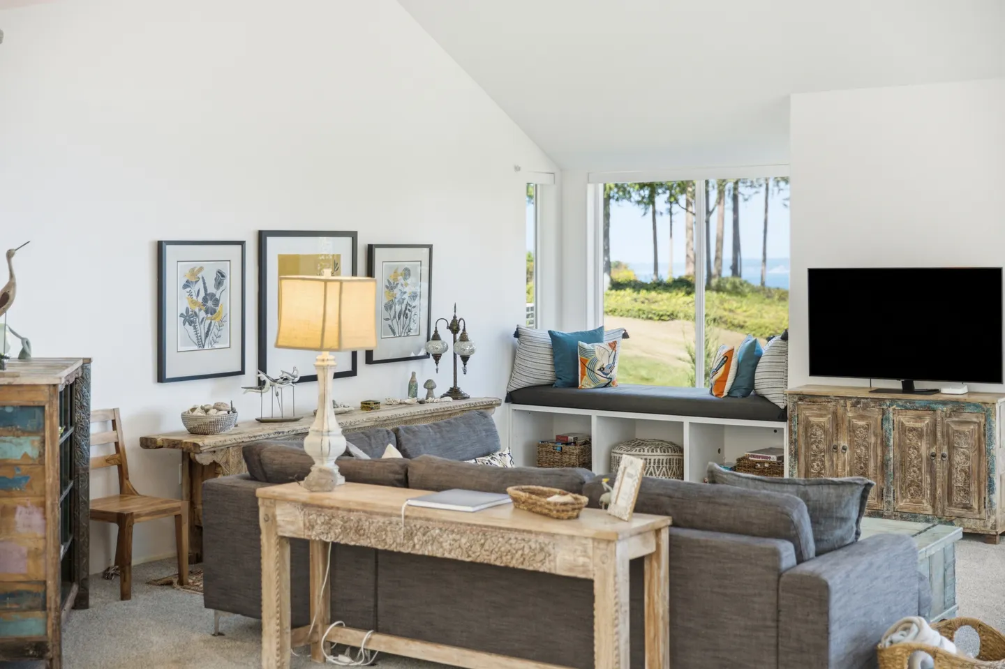 Living room with gray sofa, console table, window bench seat, television on a wooden cabinet, and ocean view through the window.