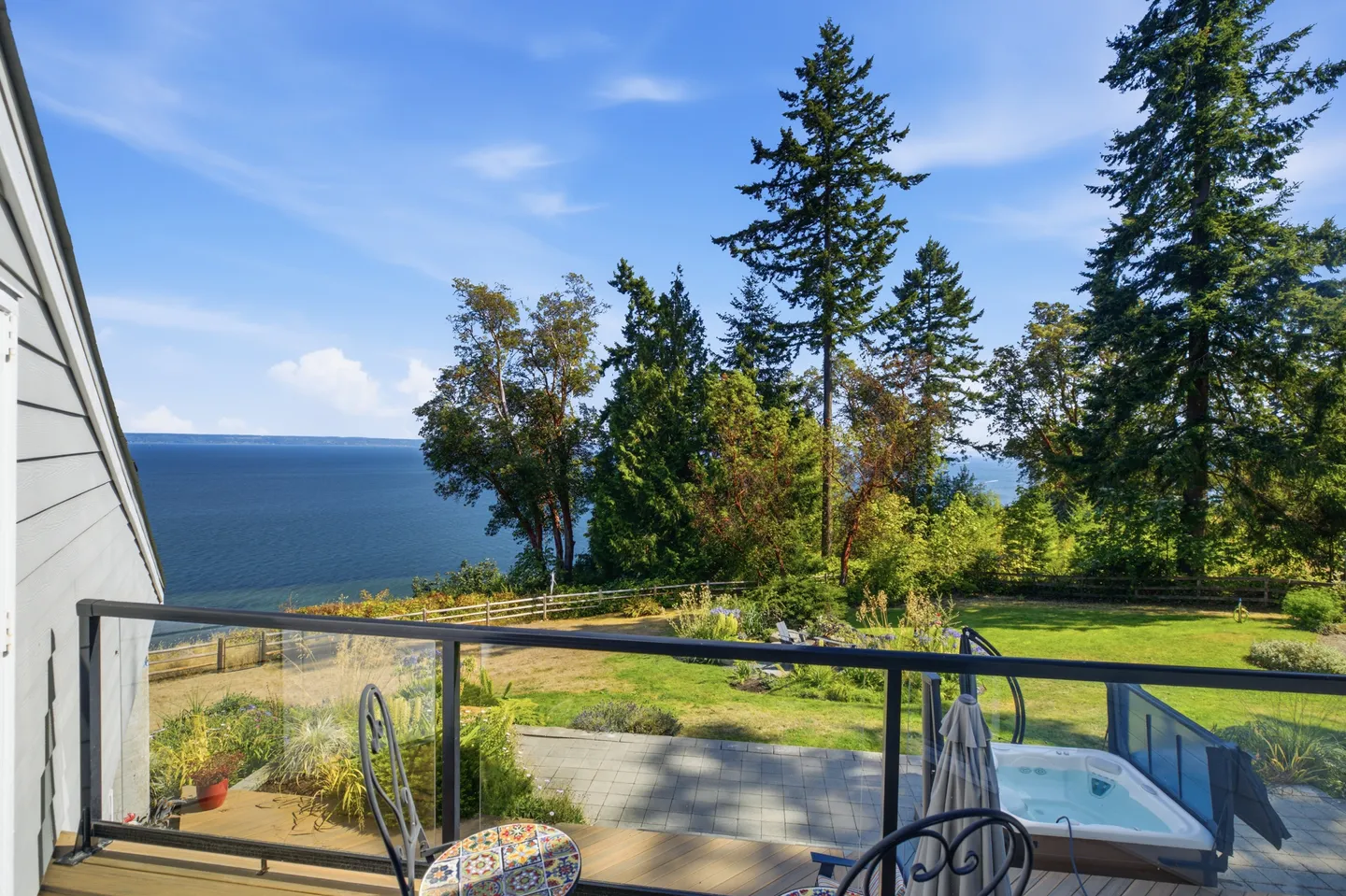 Elevated view from an upper deck overlooking a backyard with a hot tub, patio, and wide ocean view under a blue sky.