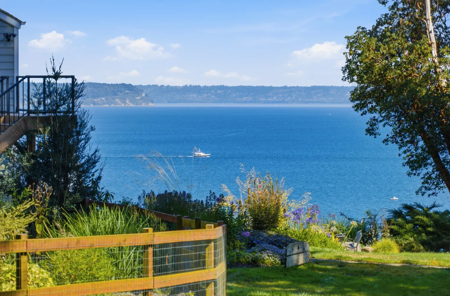 Elevated view of a large blue body of water with a boat in the distance, seen from a green lawn with a wooden fence and flowering garden.