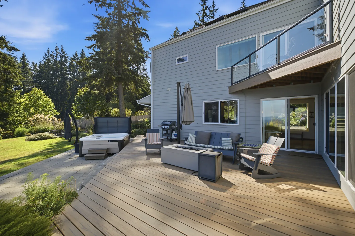 Spacious outdoor wooden deck with a fire pit table, outdoor seating, hot tub, and barbecue grill behind a two-story home.