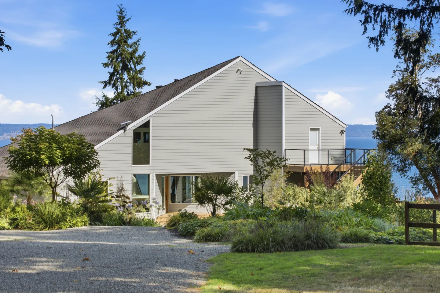 Exterior view of a multi-level home with grey siding, gravel driveway, landscaped gardens, and a view of the water in the distance.