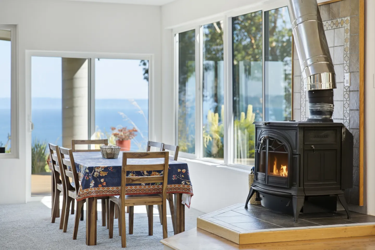 Dining area with a wood-burning stove on a raised hearth next to large windows overlooking the ocean.