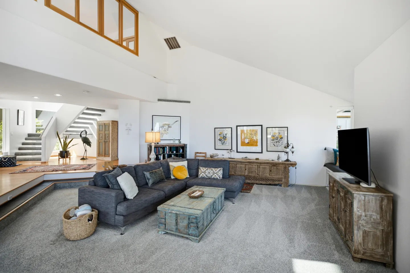 Sunlit sunken living room with high vaulted ceilings, carpet, a grey sectional sofa, coffee table trunk, TV, and stairs leading up to an adjoining level.