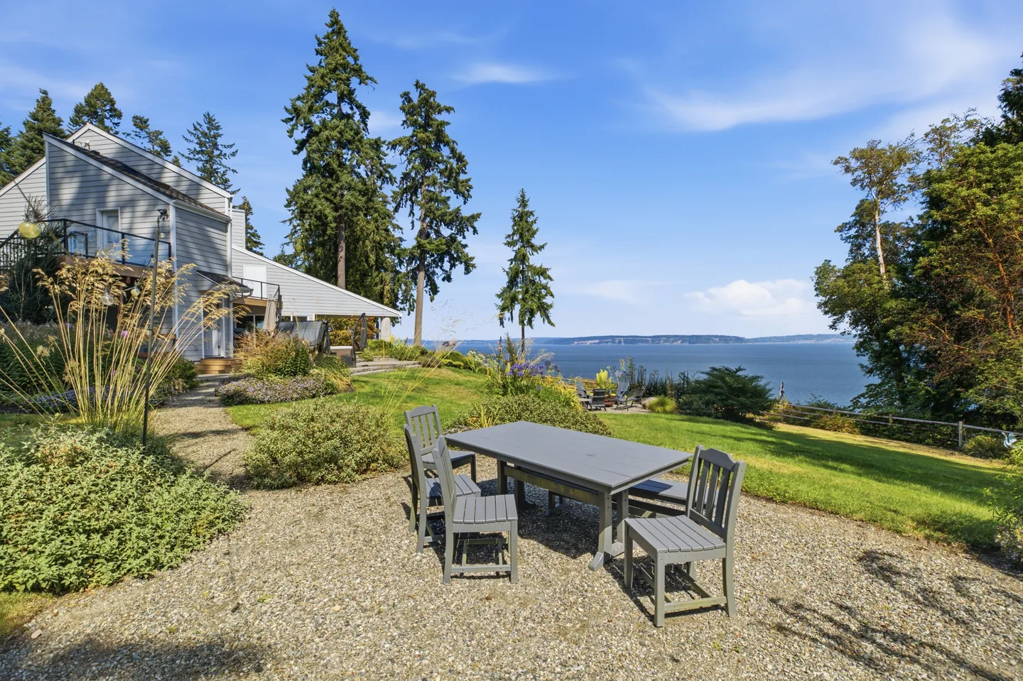 Outdoor gravel patio with a dining table and chairs overlooking a lawn, trees, and a large body of water under a blue sky.