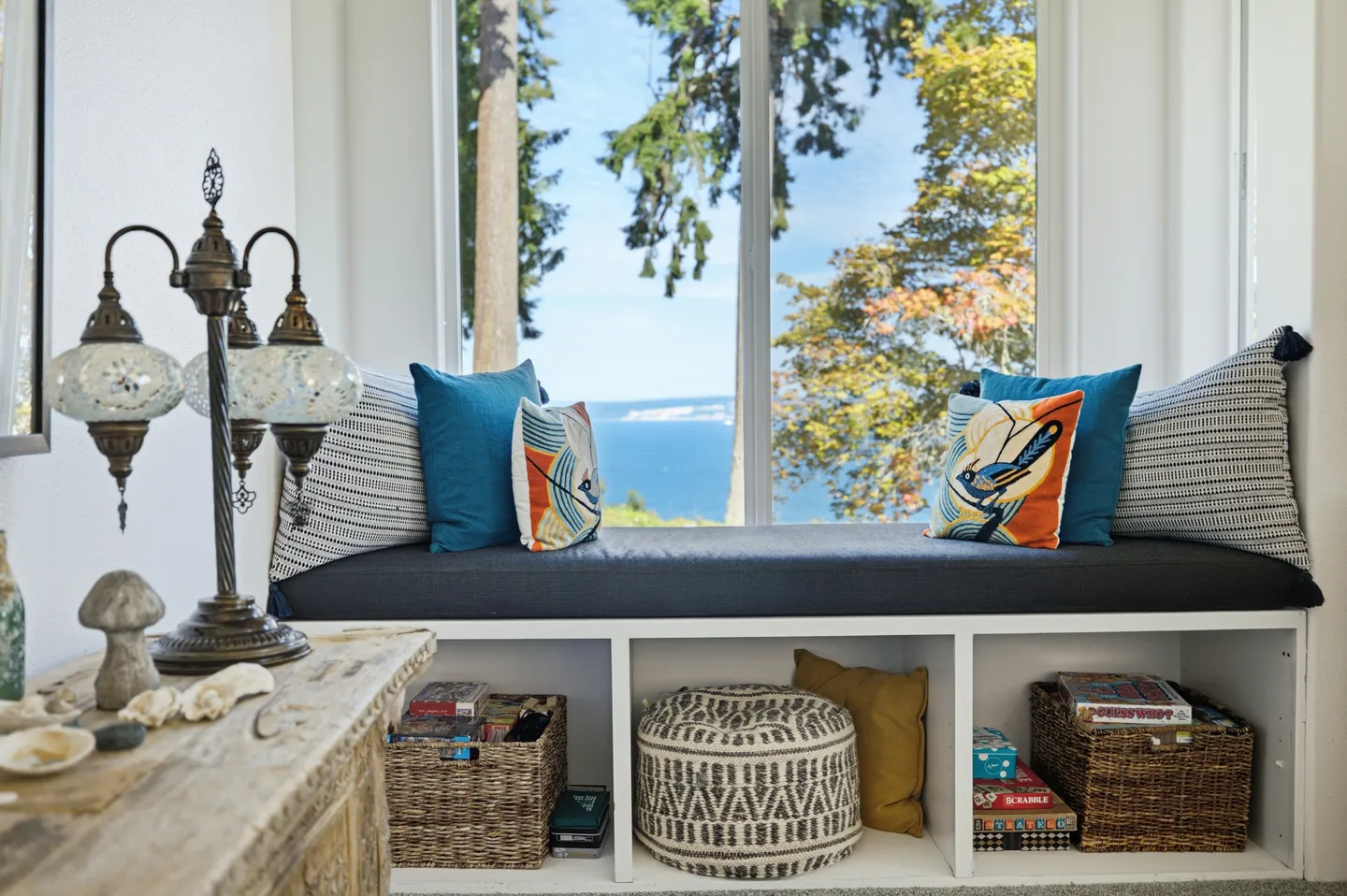 Cushioned window seat with accent pillows framing a view of trees and open water through large windows.