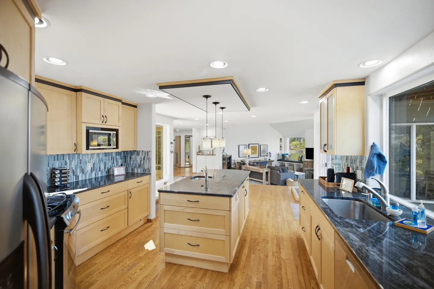 Spacious kitchen with light wood cabinetry, dark polished countertops, a central island, and wood floors looking toward an open living area.