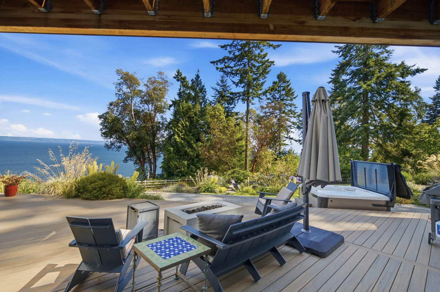 Outdoor wooden deck overlooking the water with a hot tub, fire pit table, and lounge seating.