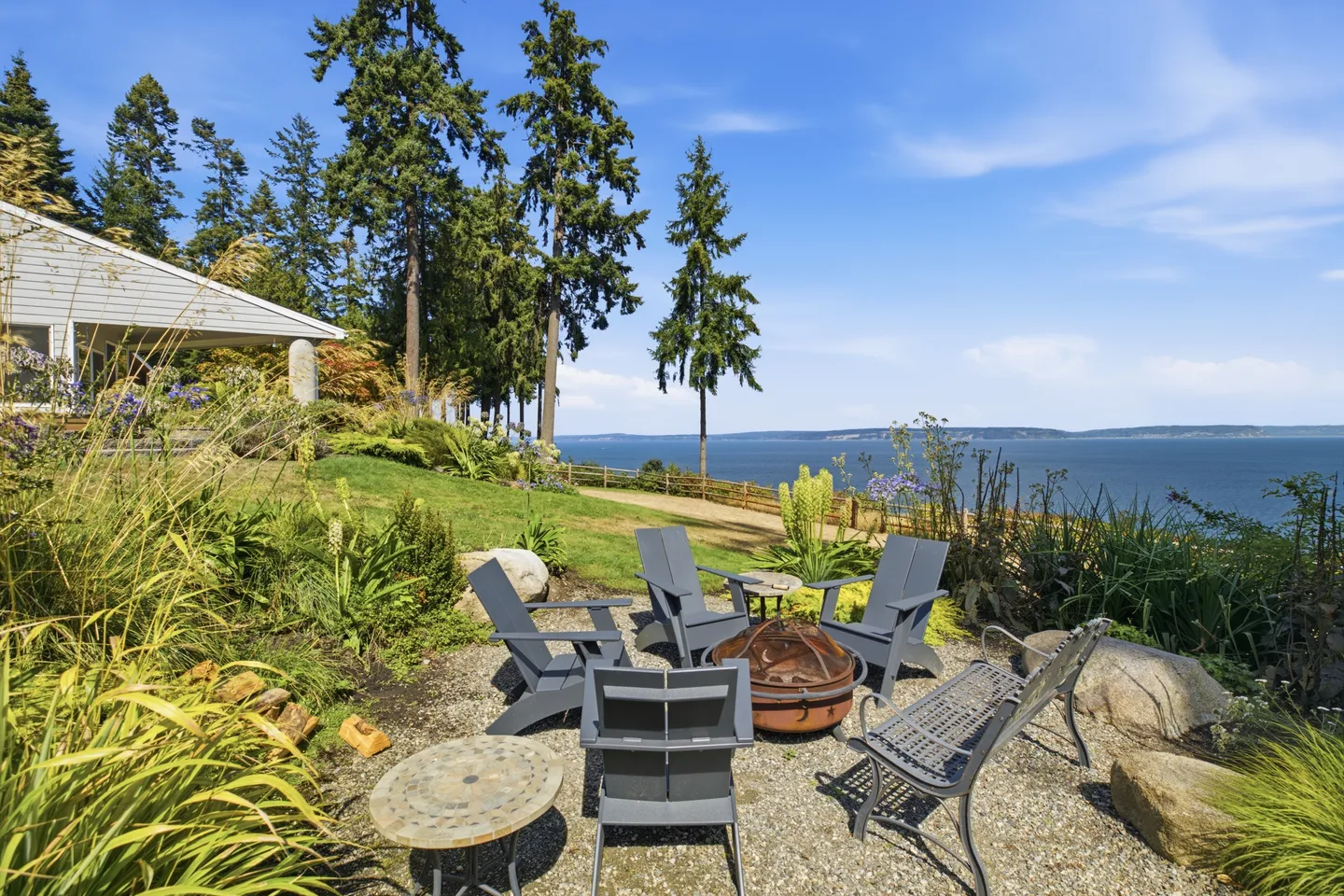 Outdoor gravel patio with a fire pit, Adirondack chairs, and a scenic water view surrounded by trees and greenery.