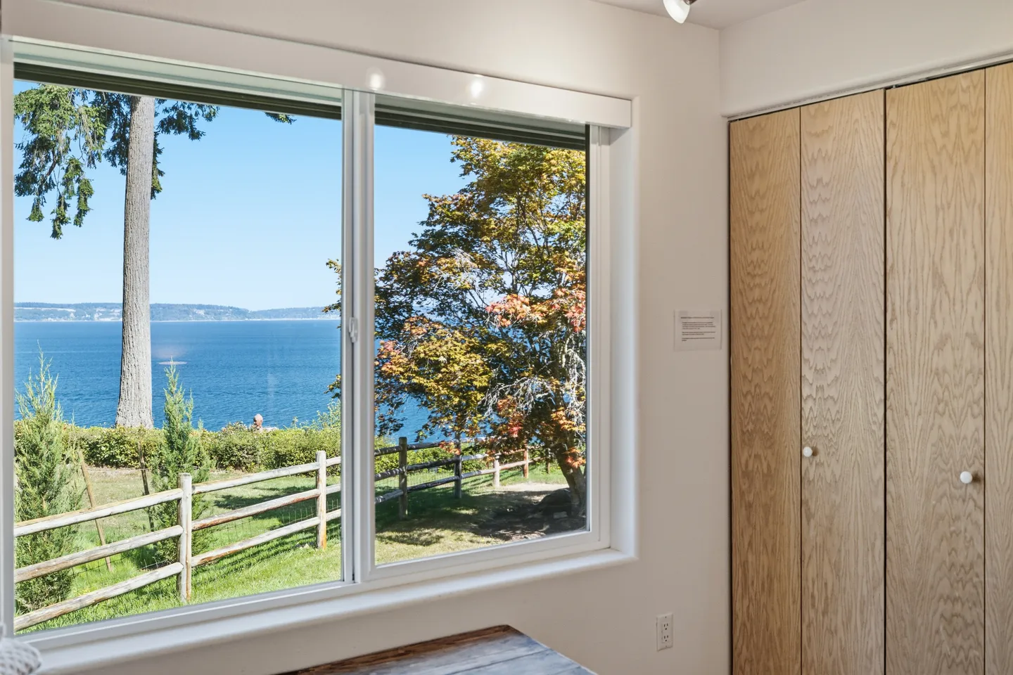 A large indoor window framing a view of a grassy yard, wooden fence, trees, and blue ocean waters under a clear sky.