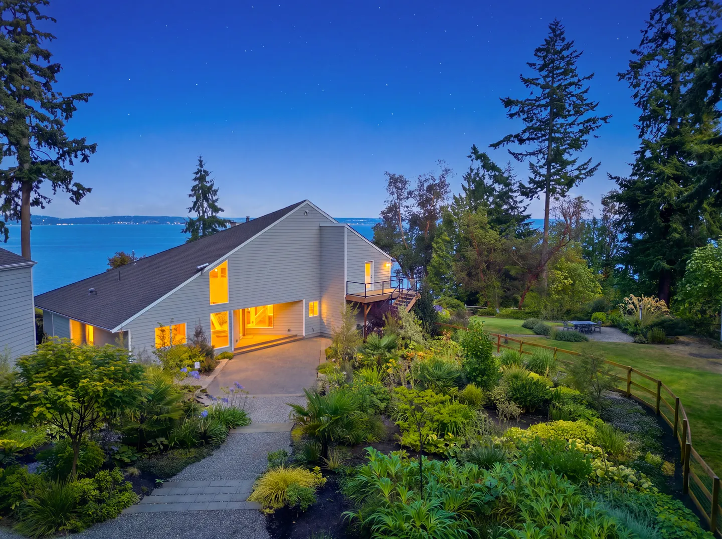 Dusk view of a contemporary home with glowing windows surrounded by lush landscaped gardens overlooking a large body of water.