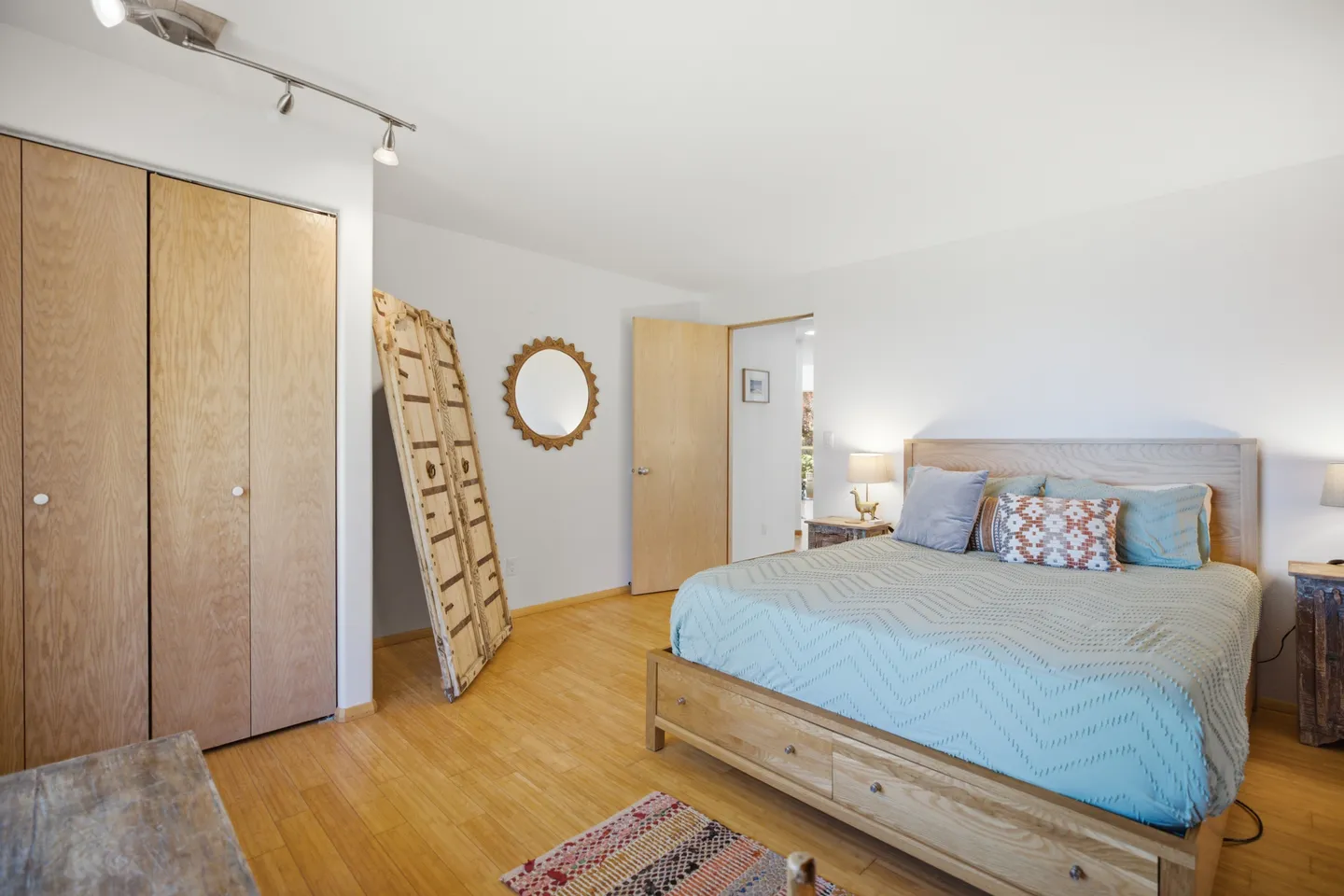 Bedroom with light wood flooring, a wooden bed frame, light blue bedding, closet doors, and decorative wall mirror.