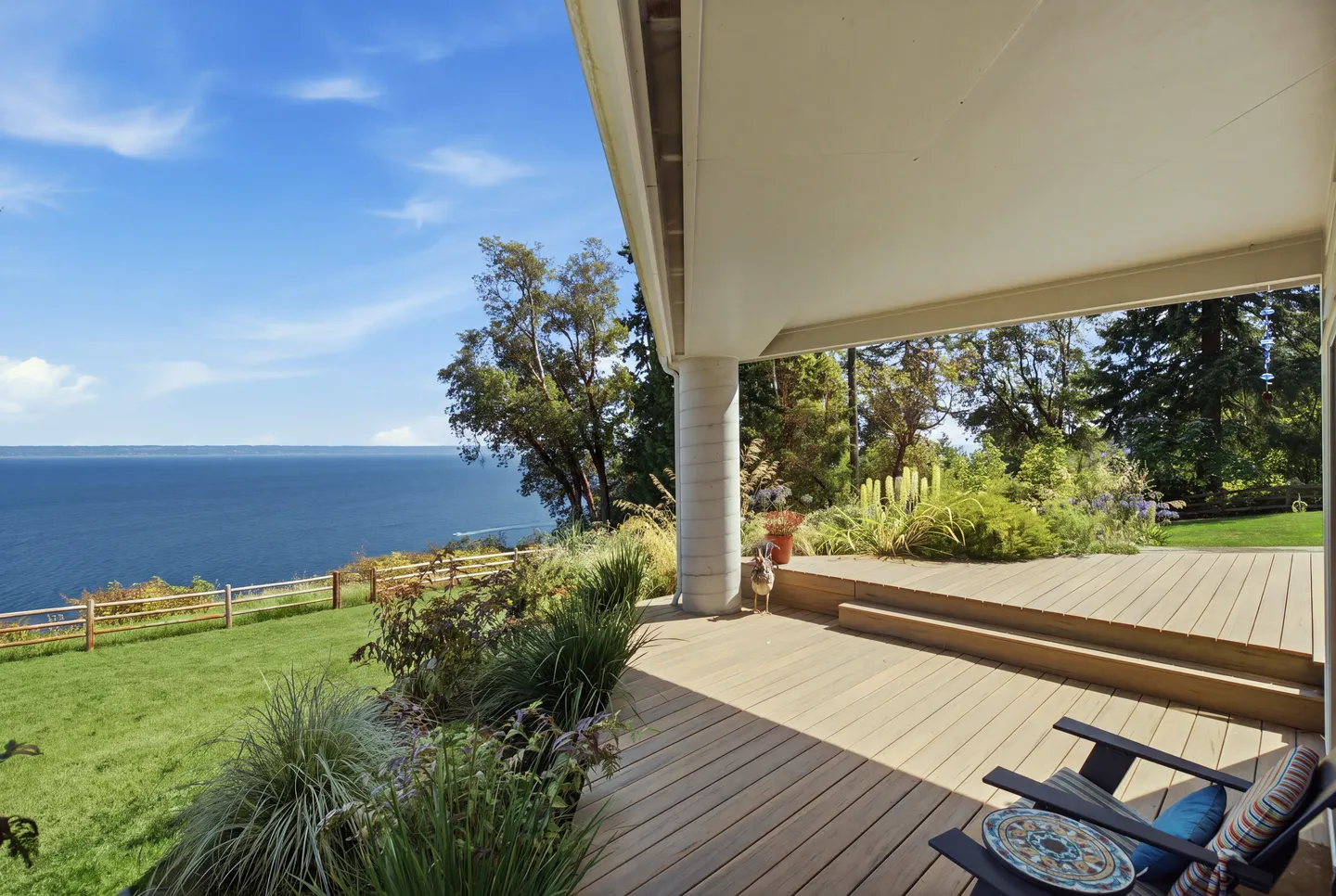 A covered outdoor wooden deck with a chair overlooking a lawn, trees, and expansive blue water under a clear sky.