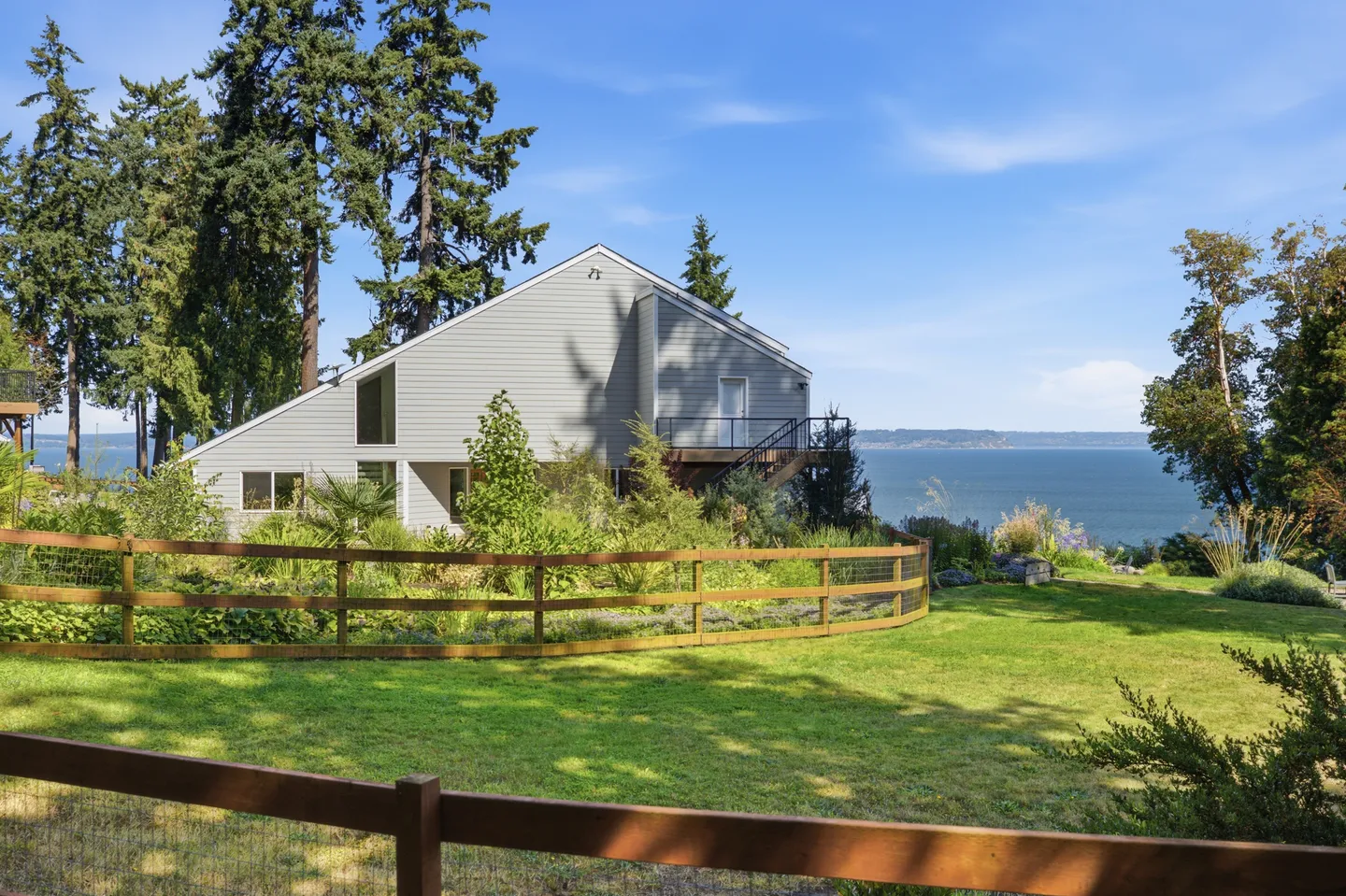 Exterior of a grey multi-story house on a grassy property overlooking a large body of water under a clear blue sky.
