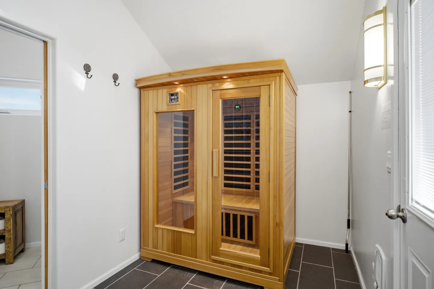 A wooden freestanding sauna cabin situated in a room with white walls and dark tiled flooring.