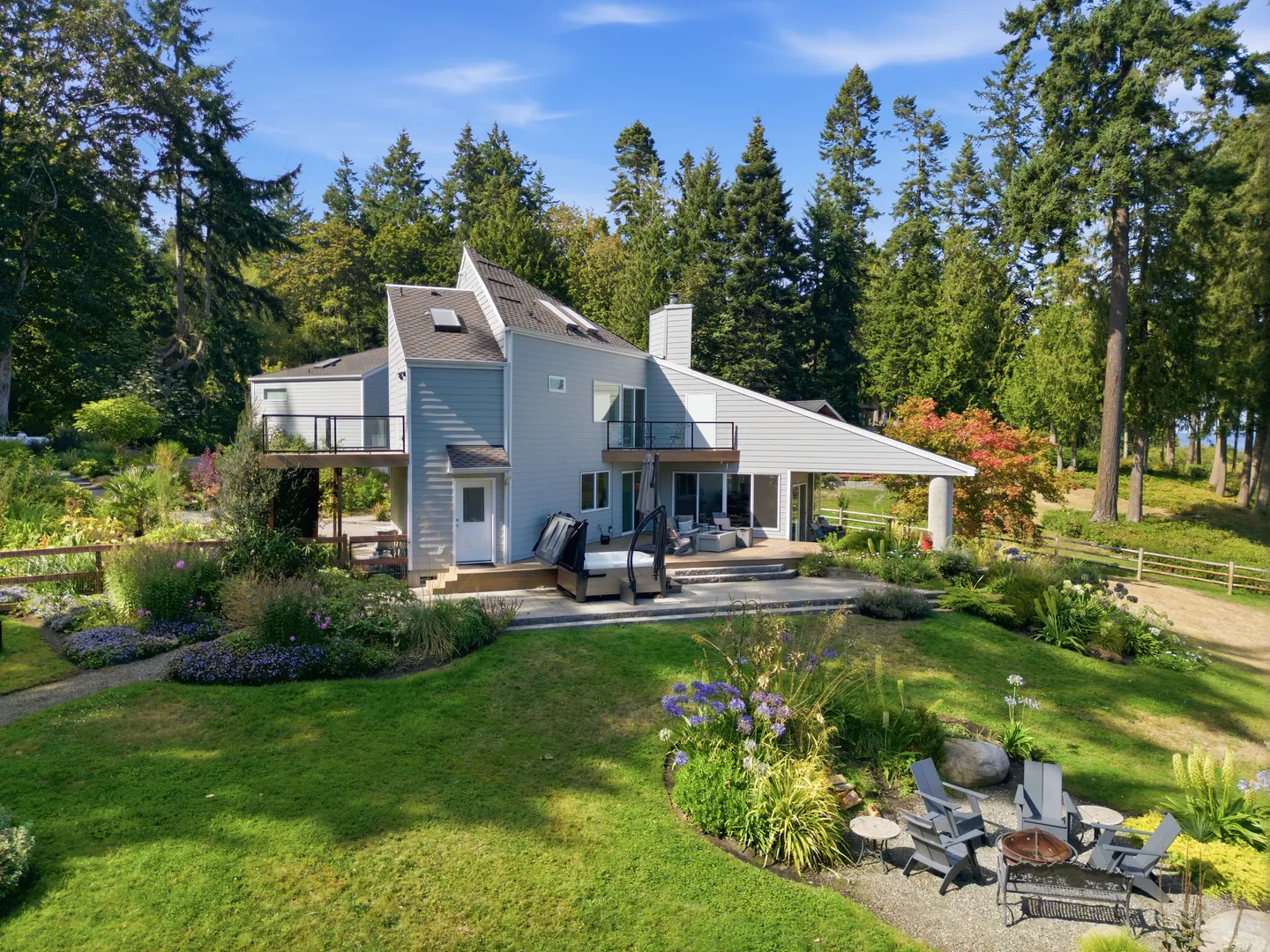 Exterior view of a multi-story home with light blue siding, surrounded by a green lawn, landscaping, a hot tub, and a fire pit area.