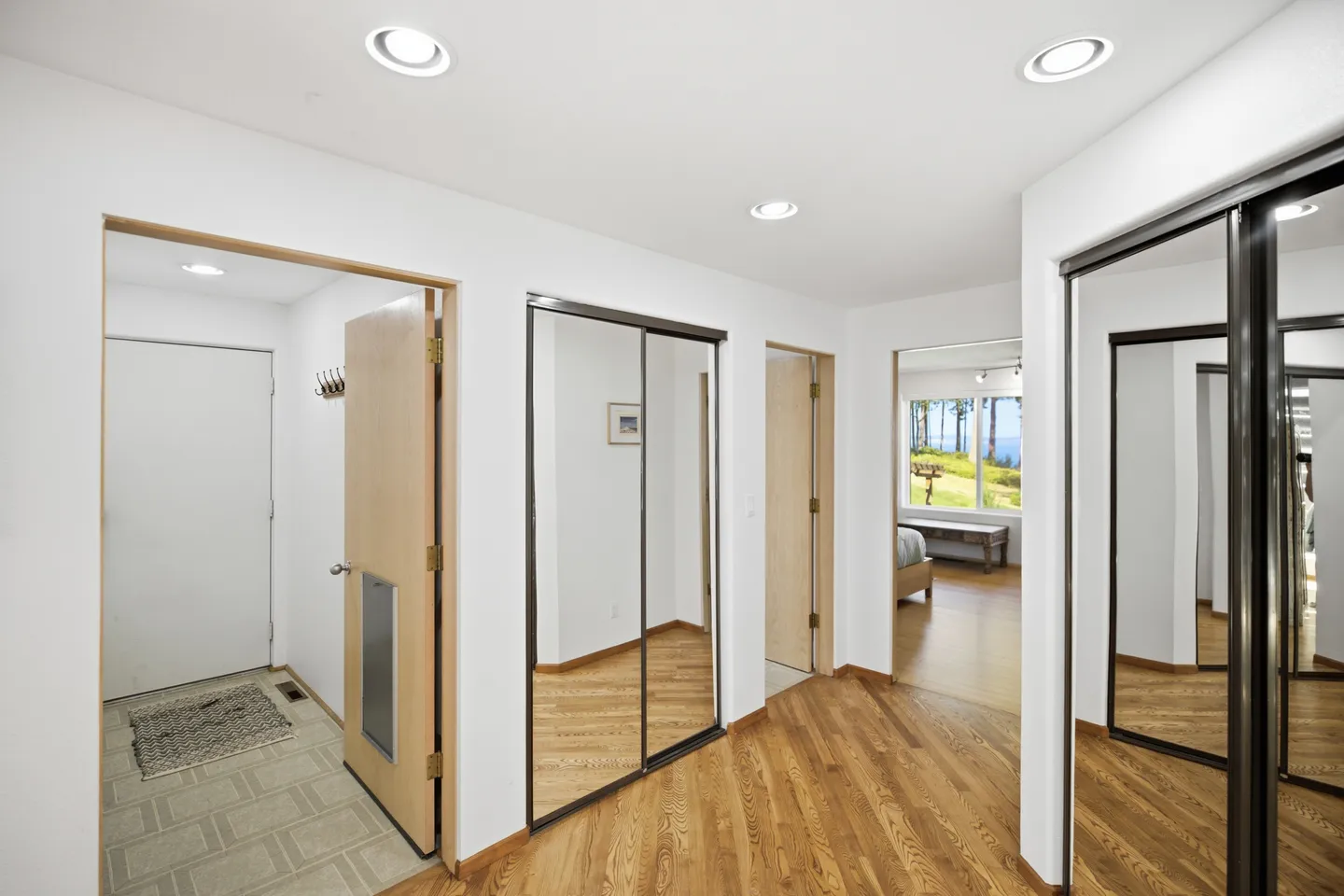 Hallway with wood floors, mirrored closet doors, and recessed ceiling lights leading toward an entryway and bedroom.