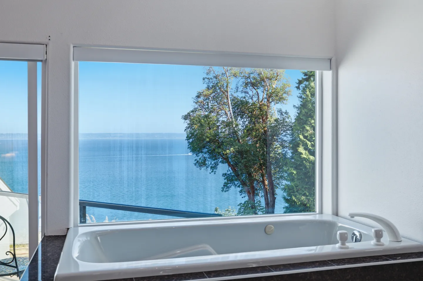 A built-in soaking tub positioned below a large picture window offering a view of open water and trees on a sunny day.
