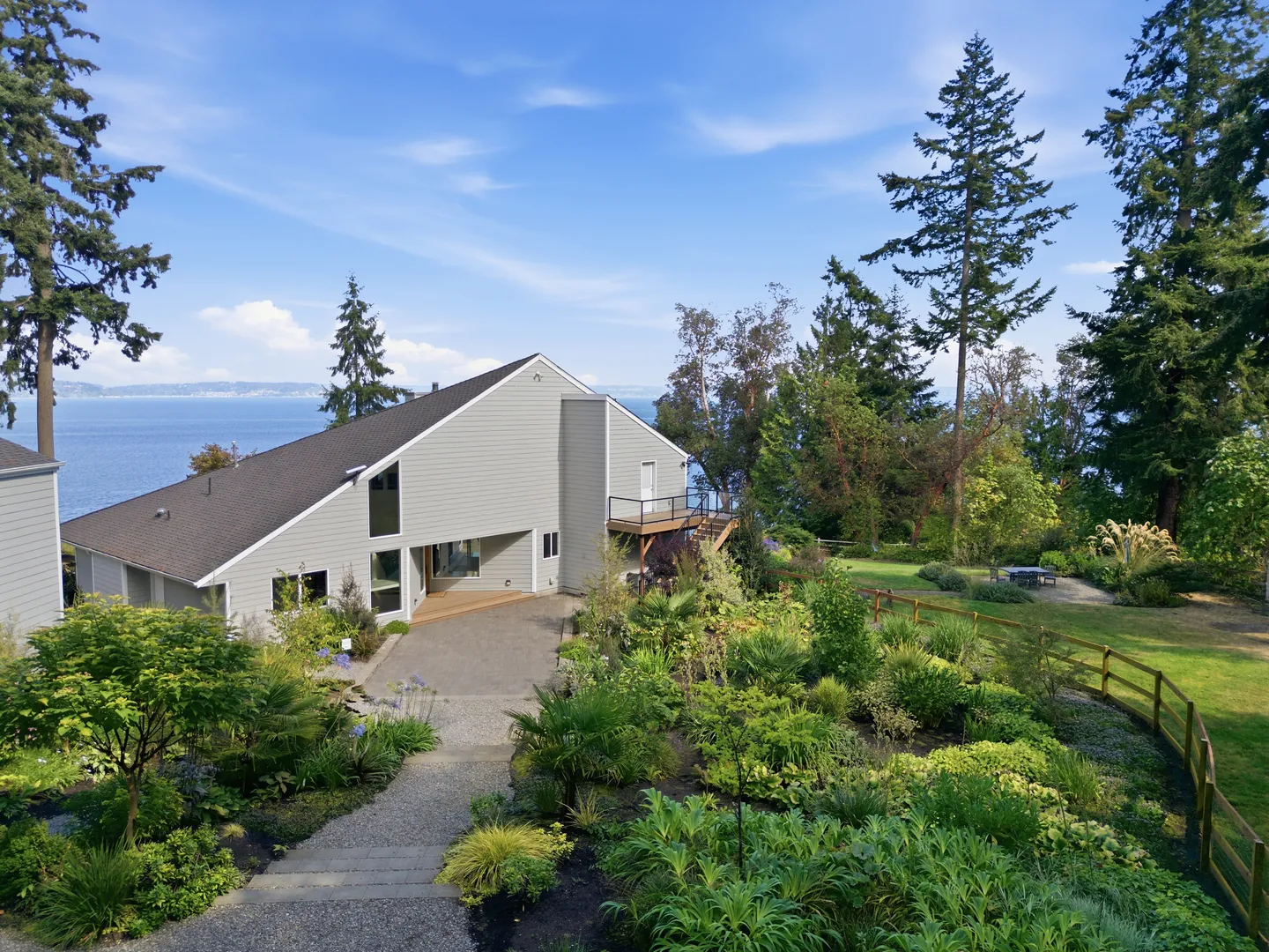 Elevated view of a home exterior with light siding, surrounded by lush landscaped gardens, tall trees, and a view of the water.