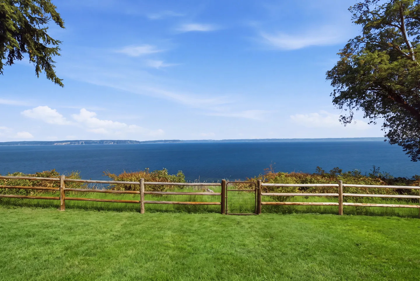 A wide view of a large body of water seen over a green lawn and wooden fence under a blue sky.