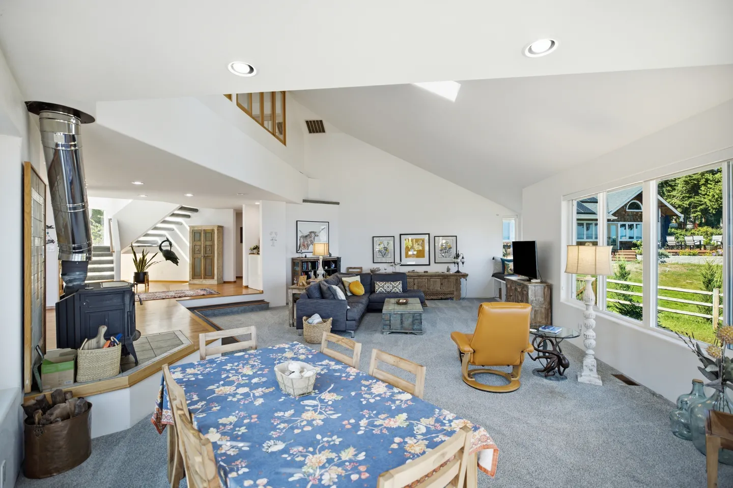 Open-concept living and dining area with vaulted ceiling, wood-burning stove, dining table, sofa, and large windows looking outside.