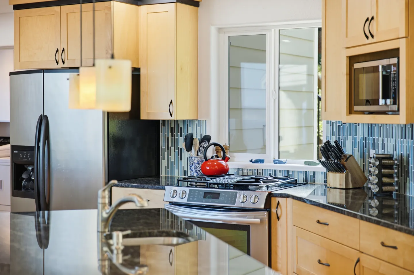 Close-up view of a kitchen featuring light wood cabinets, dark granite countertops, stainless steel appliances, and a blue tile backsplash.