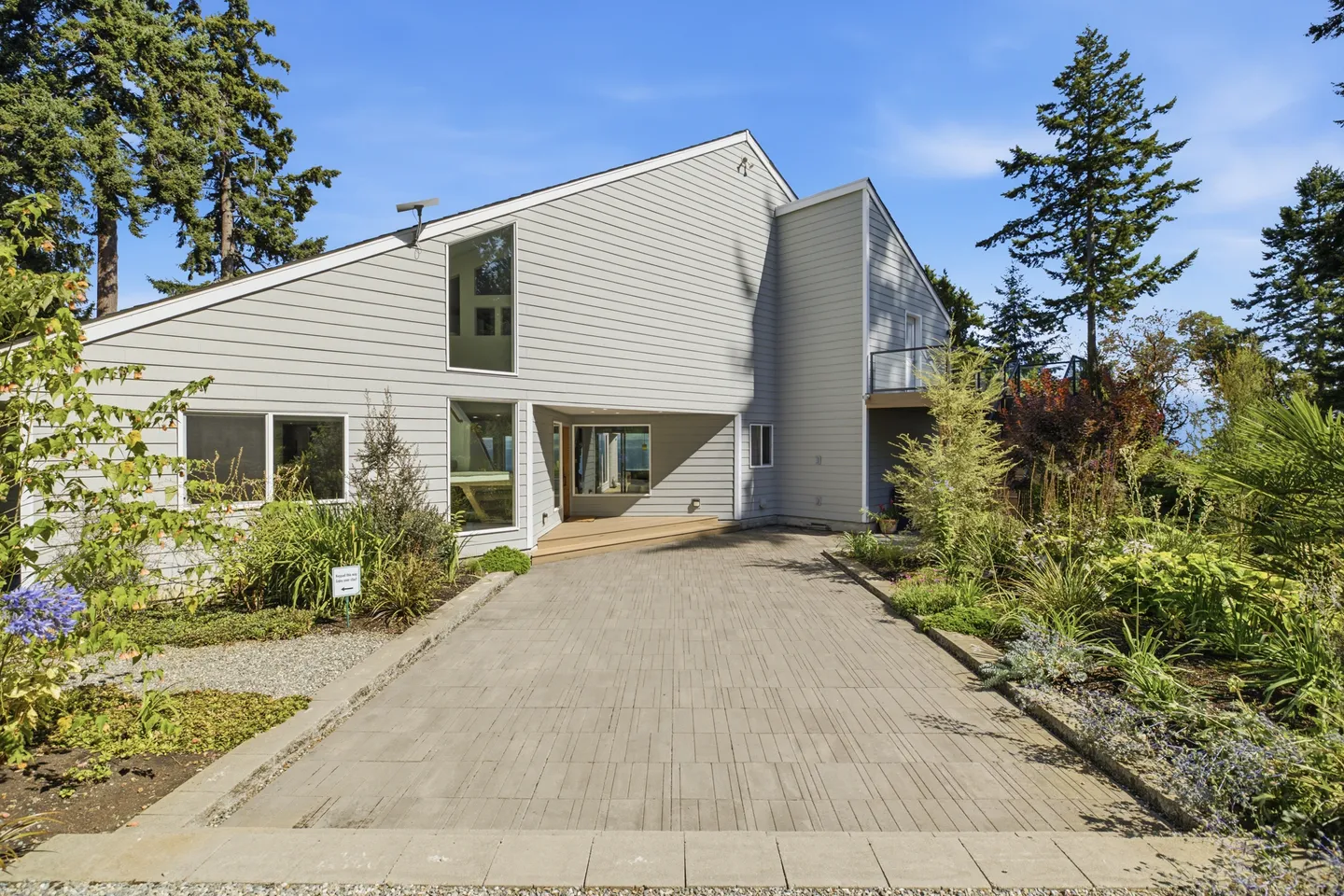 Exterior front view of a two-story home with light grey siding and a wide paved driveway leading to the entrance under a clear blue sky.
