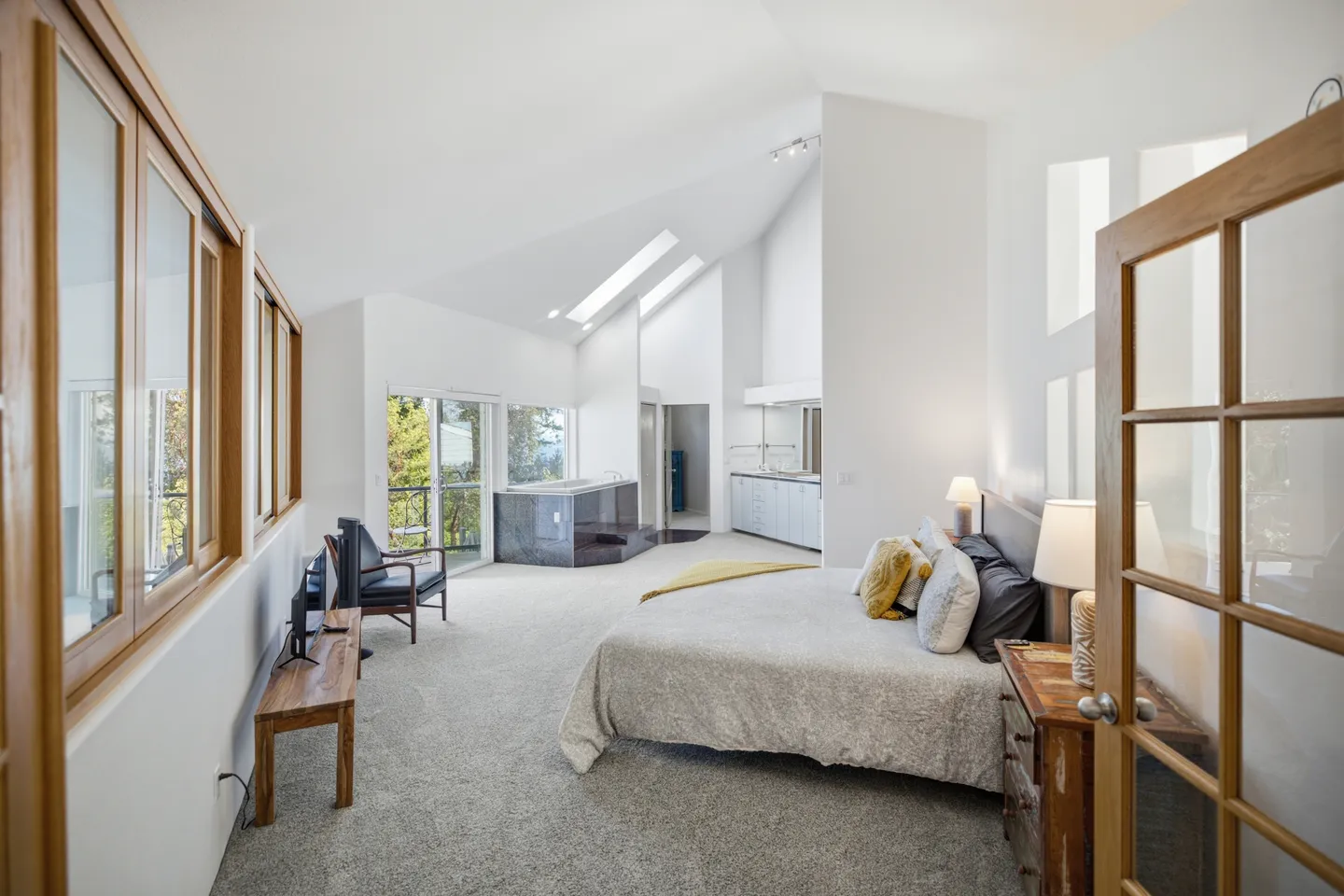 Spacious bedroom suite with carpeted flooring, vaulted ceiling with skylights, a bed, and an elevated corner soaking tub in the background.