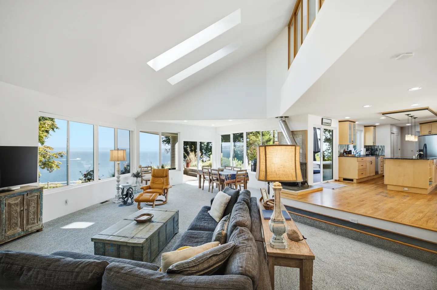 Open-concept living room with vaulted ceilings, skylights, large windows overlooking water, a sectional couch, and a view into the dining area and kitchen.