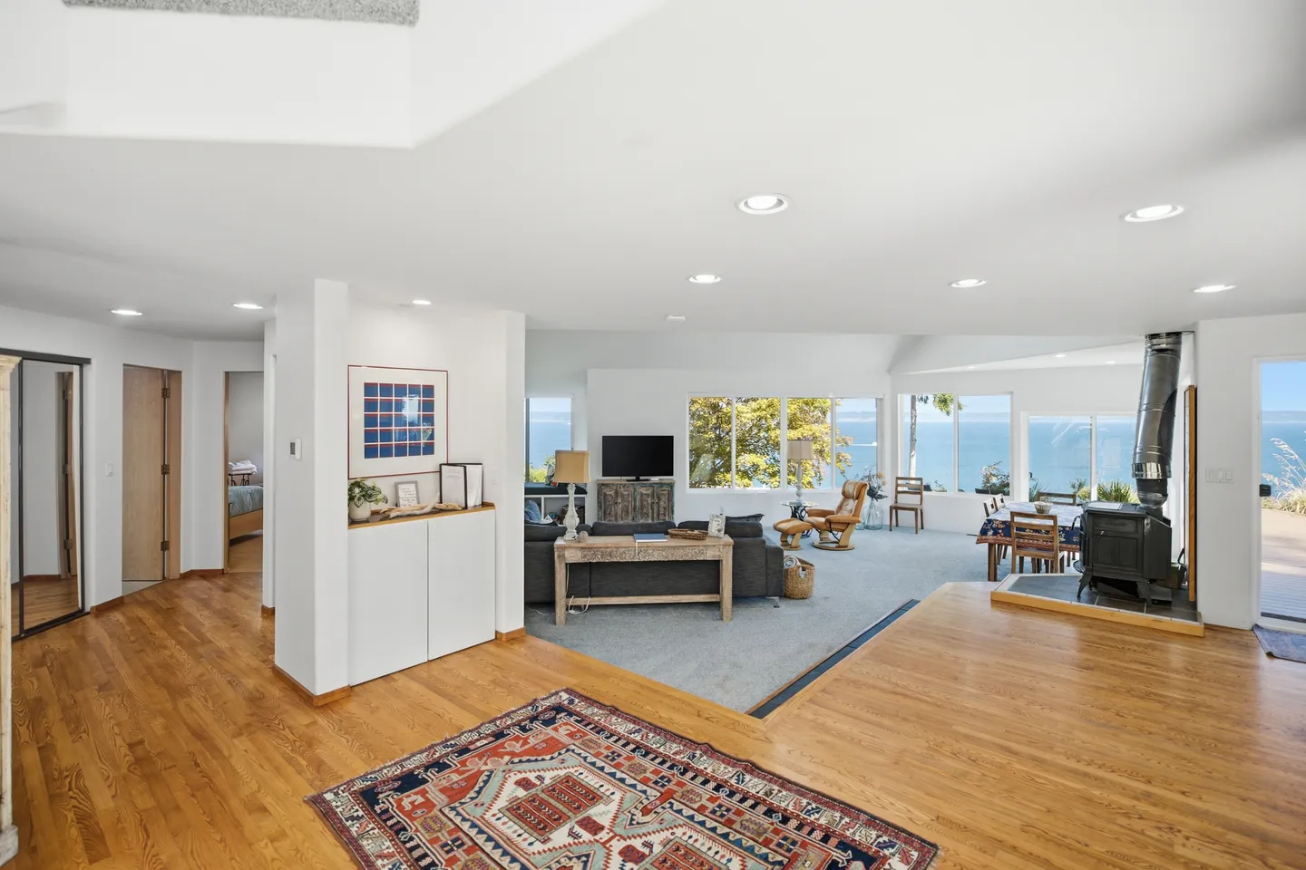Open-concept living room and dining area with hardwood floors, a sunken carpeted sitting space, a wood stove, and large windows overlooking the water.