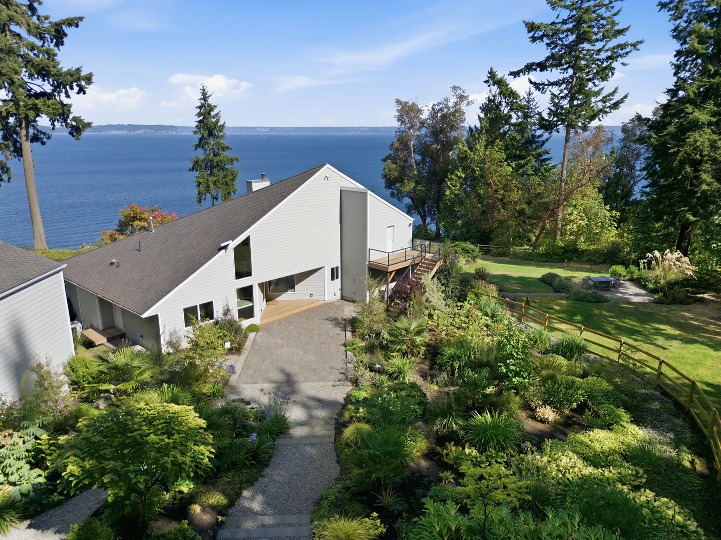 Aerial view of a contemporary light-gray house with landscaped gardens and paved walkways overlooking a vast body of water.