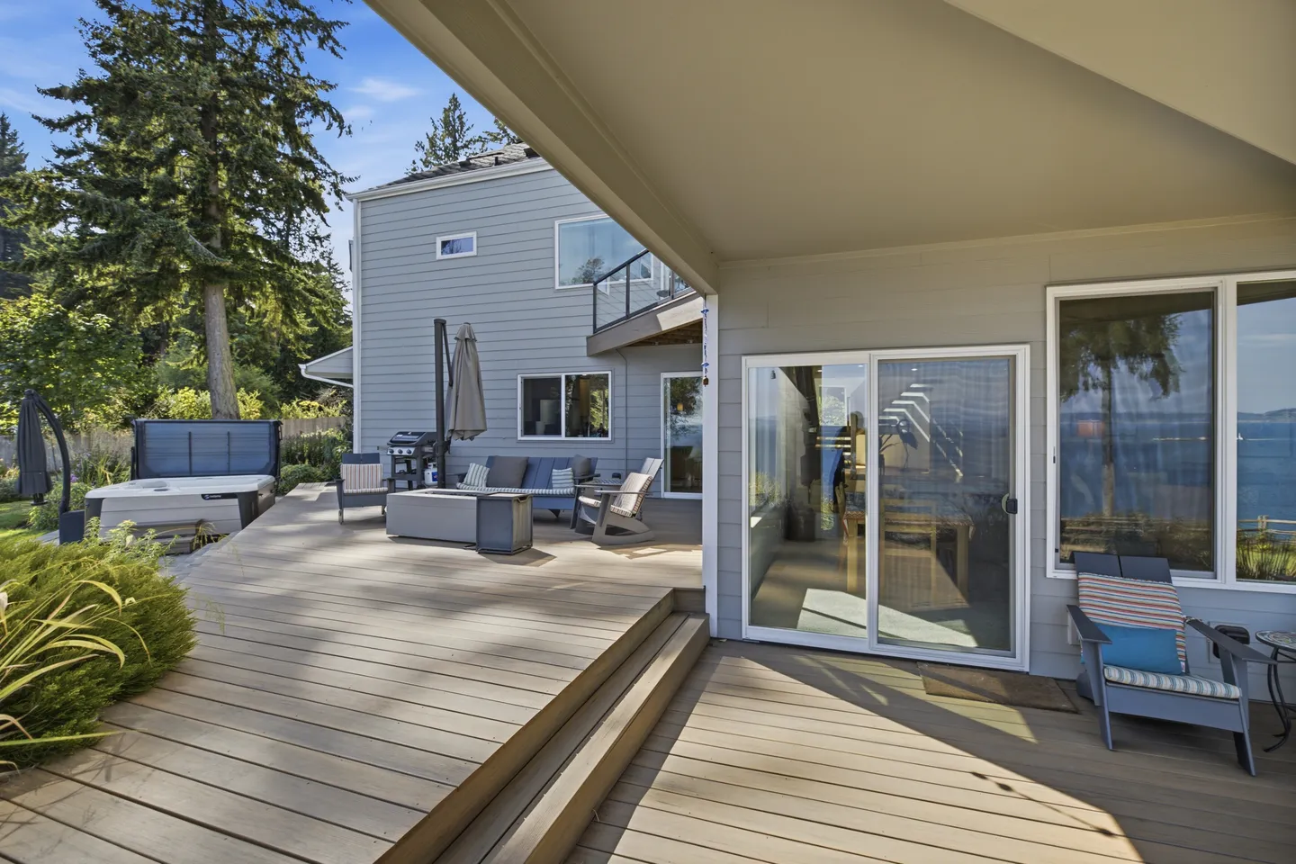 A wide multi-level wooden deck behind a two-story home, featuring patio seating, a fire table, a hot tub, and a partial view of the water.