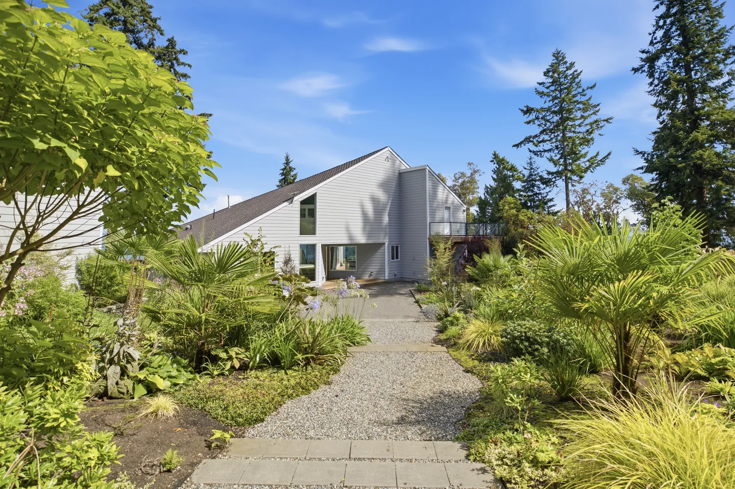 A gravel and stone walkway flanked by palm trees and lush landscaping leads toward a modern gray home under a blue sky.