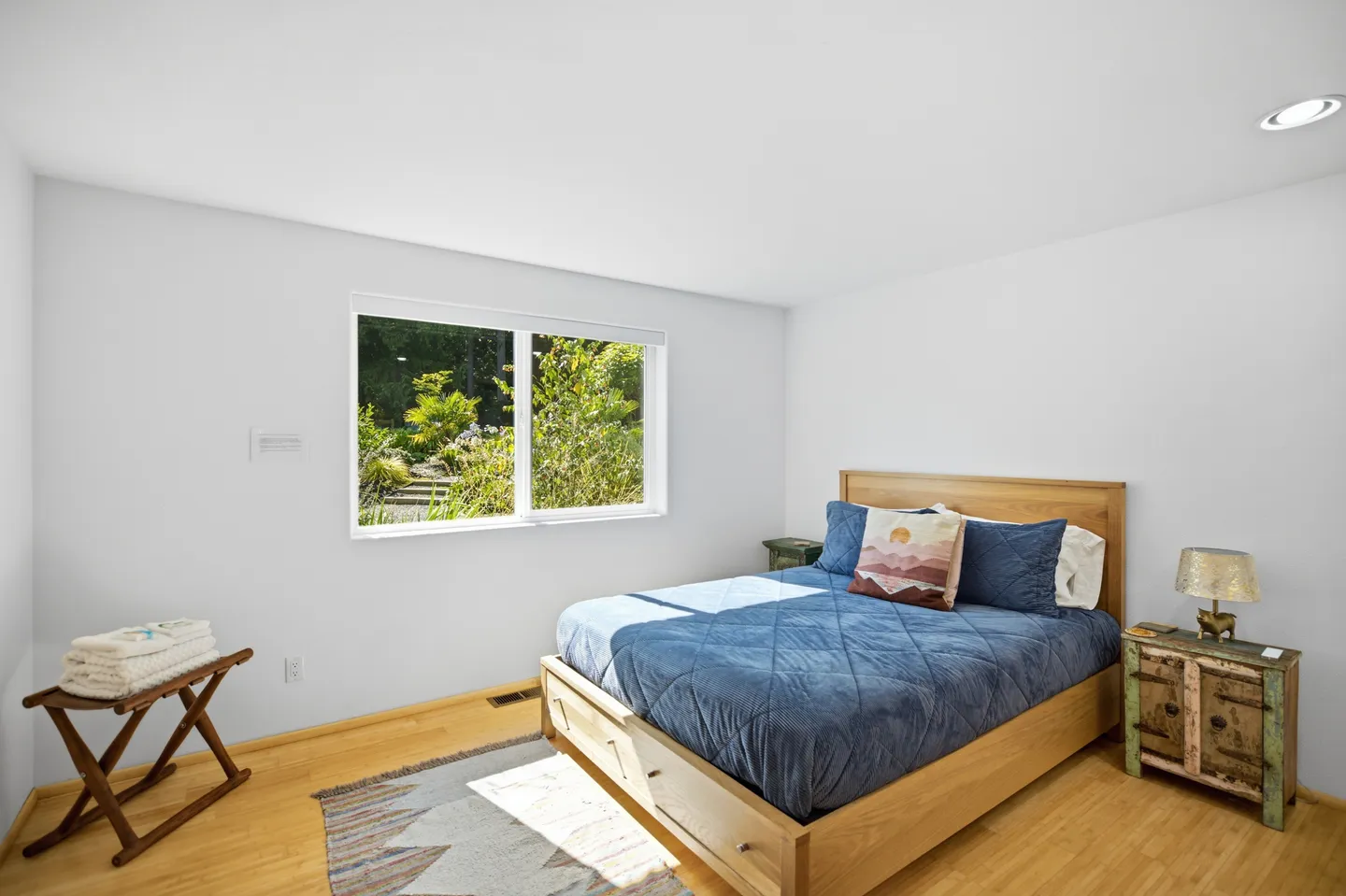 Bedroom with wooden platform bed, blue bedding, light wood floors, and a window overlooking lush green foliage.