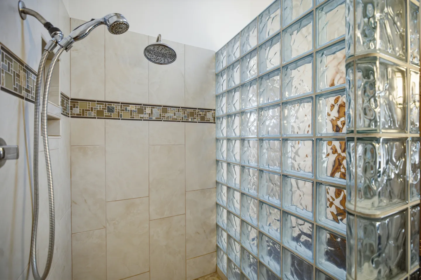 Tiled walk-in shower with an overhead showerhead, handheld sprayer, decorative tile border, and a glass block wall.