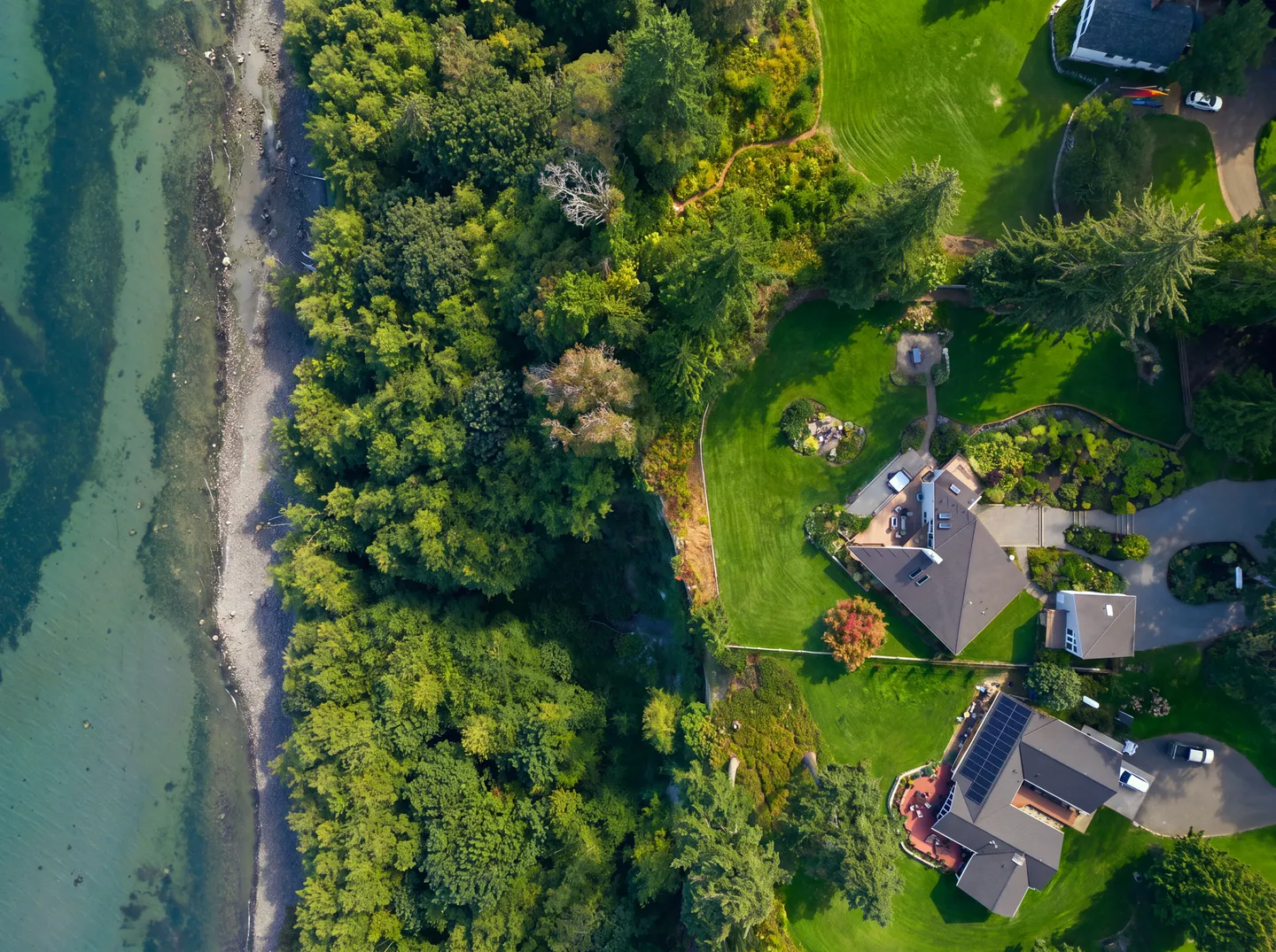 Aerial top-down view of coastal properties bordered by a green forest and a rocky beach next to clear water.