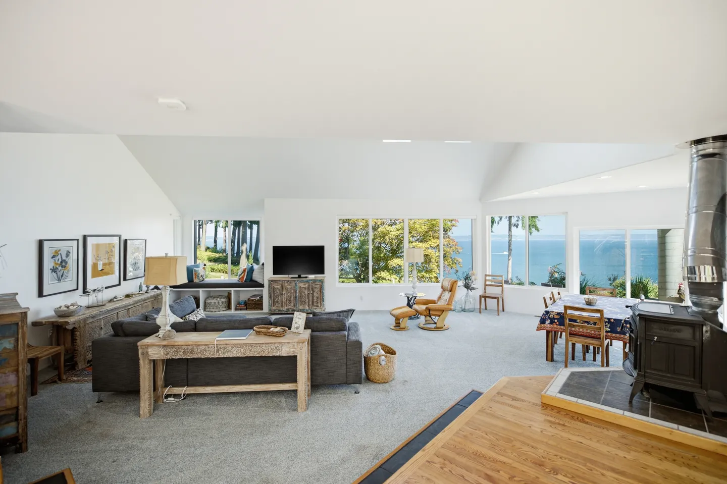 Open-concept living room and dining area with carpeted floors, large windows overlooking water, a wood-burning stove, and vaulted ceiling.