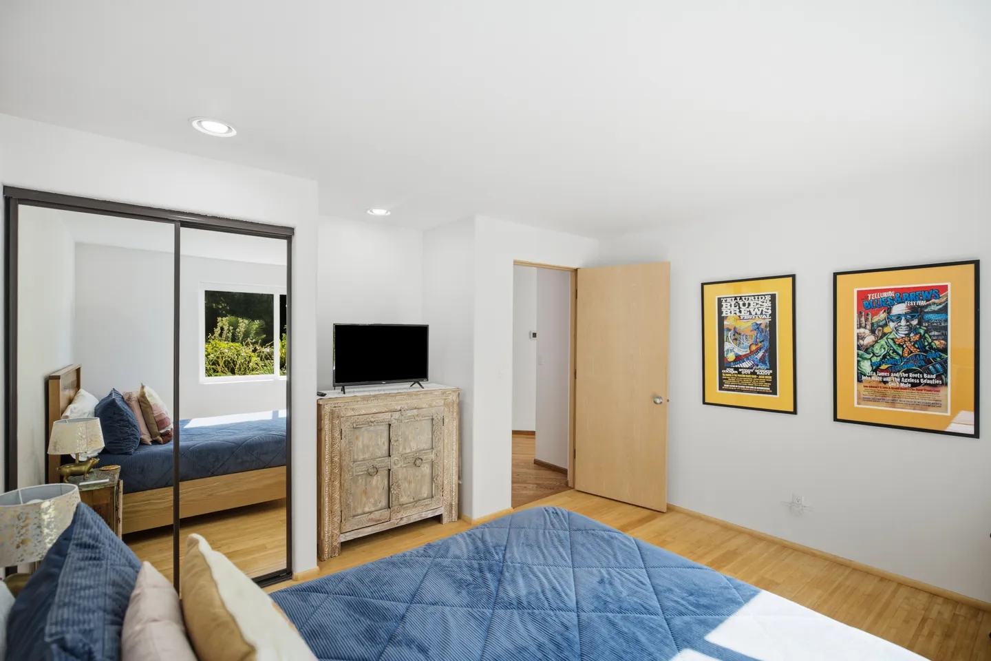 Bedroom with hardwood floors, a bed with blue bedding, mirrored closet doors, a wooden cabinet with a TV, and framed posters on the wall.