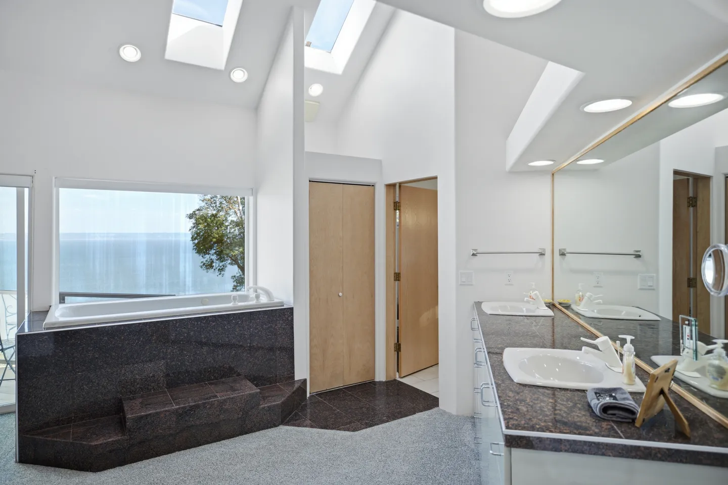 Primary bathroom featuring a deep soaking tub surrounded by dark polished stone next to a large picture window overlooking the water.