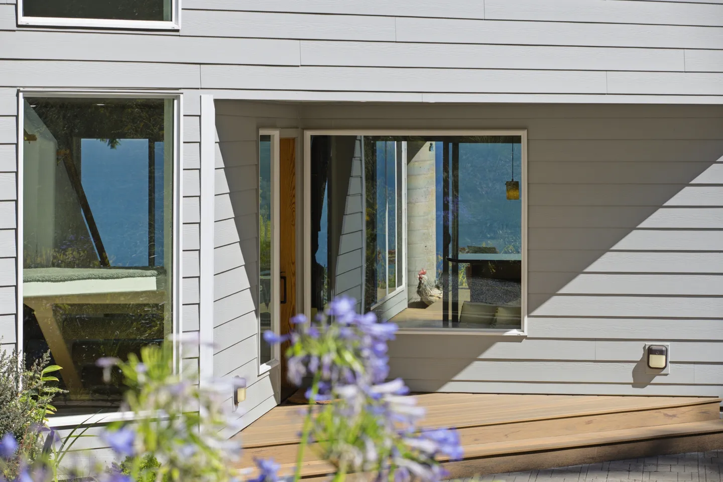 Exterior view of a home entrance with light horizontal siding, large picture windows reflecting water, and a wooden deck.