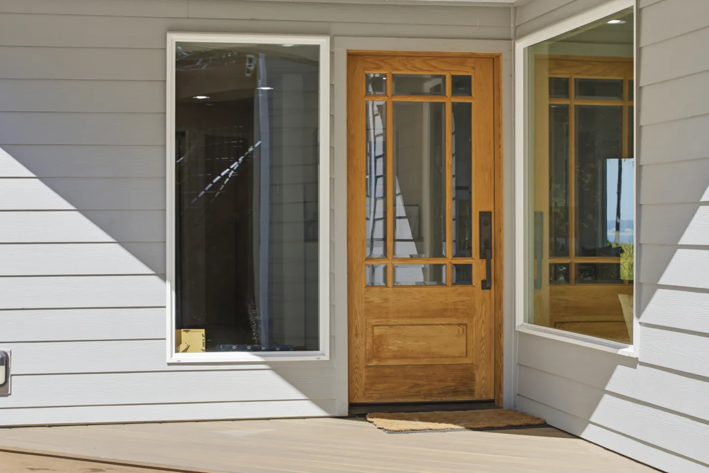 A wooden exterior door with glass window panes set into light grey siding next to large windows.
